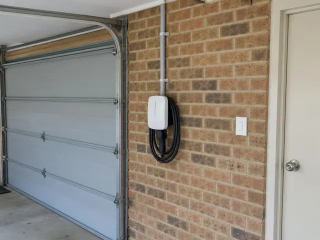 Wall-mounted EV charger on a brick garage wall beside a roller door in Marks Point